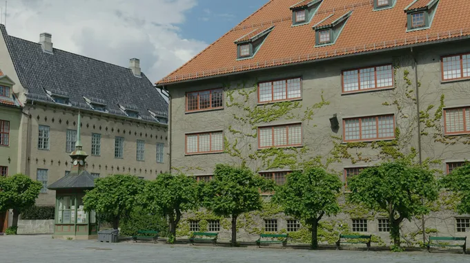 Historisk bygning med oransje takstein og grønne trær i forgrunnen. Bygningen har flere vinduer med trekarm og en grå fasade
